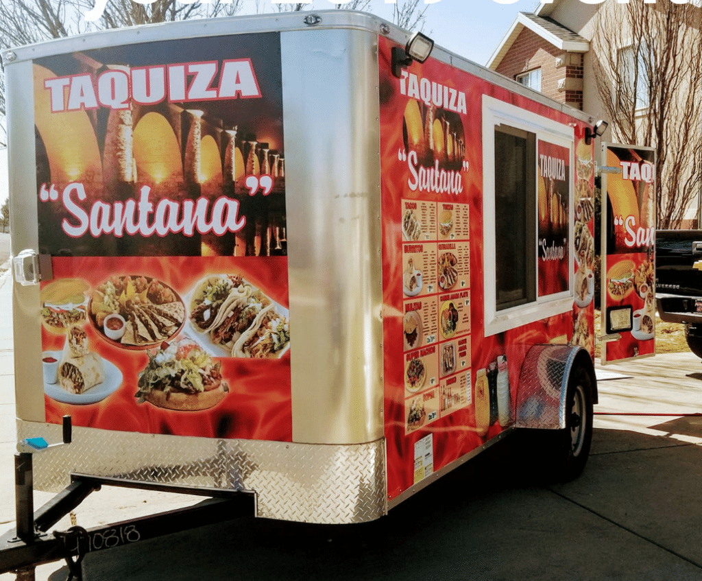 Colorful food truck named ‘Taquiza Santana’ with vibrant images of tacos, burritos, and other Mexican dishes displayed on its exterior under bright sunlight.
